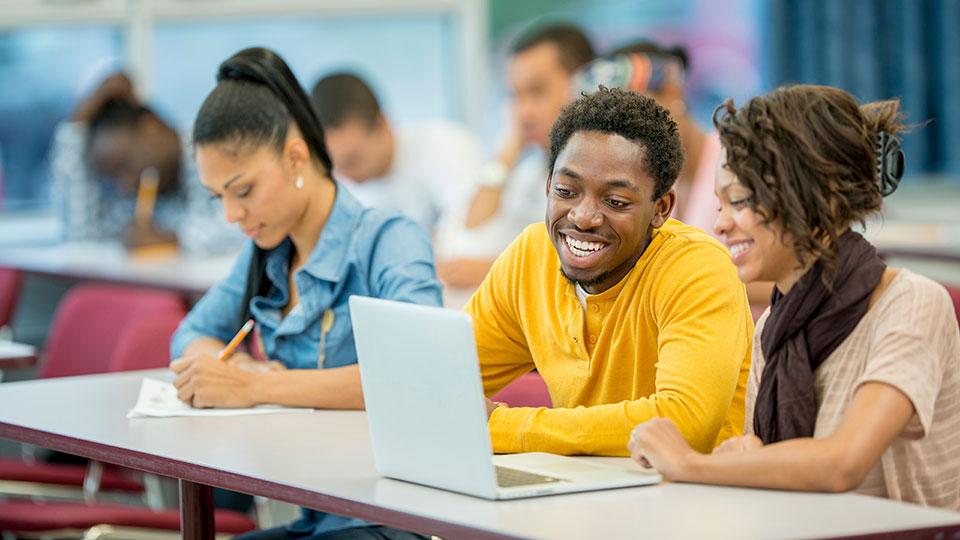 Three Black students sit at a table, with two of them working on a laptop, and the third writing on a piece of paper